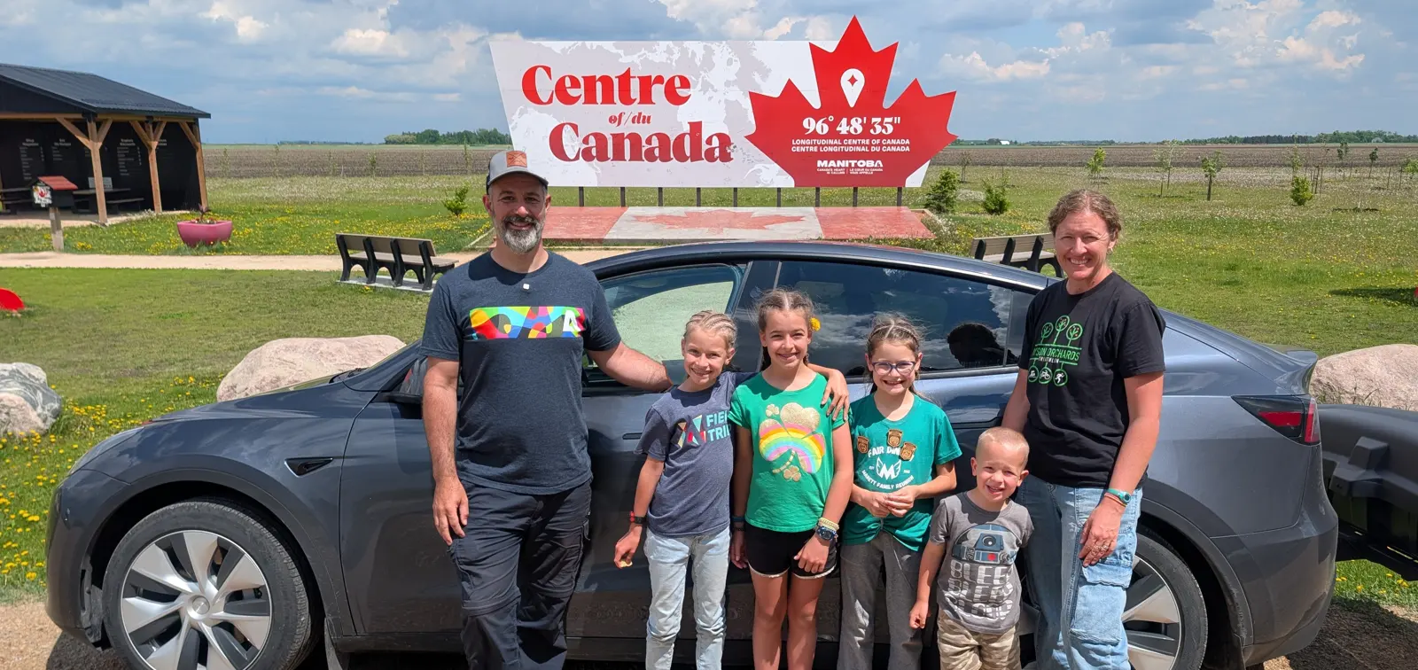 Our family of six standing in front of our Tesla Model Y at the Centre of Canada sign in Manitoba